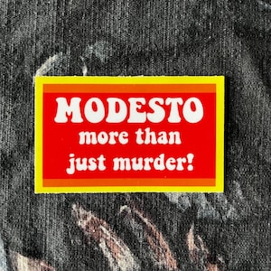 May include: A rectangular sticker with the word "MODESTO" in white, bold font on a red background. Below, the text reads "more than just murder!" in a smaller white font. The sticker has a yellow border with orange stripes.