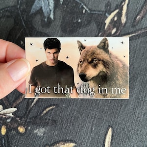Op de afbeelding: Een rechthoekige sticker met een man en een wolf, met de tekst "I got that dog in me". De man draagt een donker shirt. De wolf heeft bruine vacht. De achtergrond is lichtoranje met zwarte sterren.