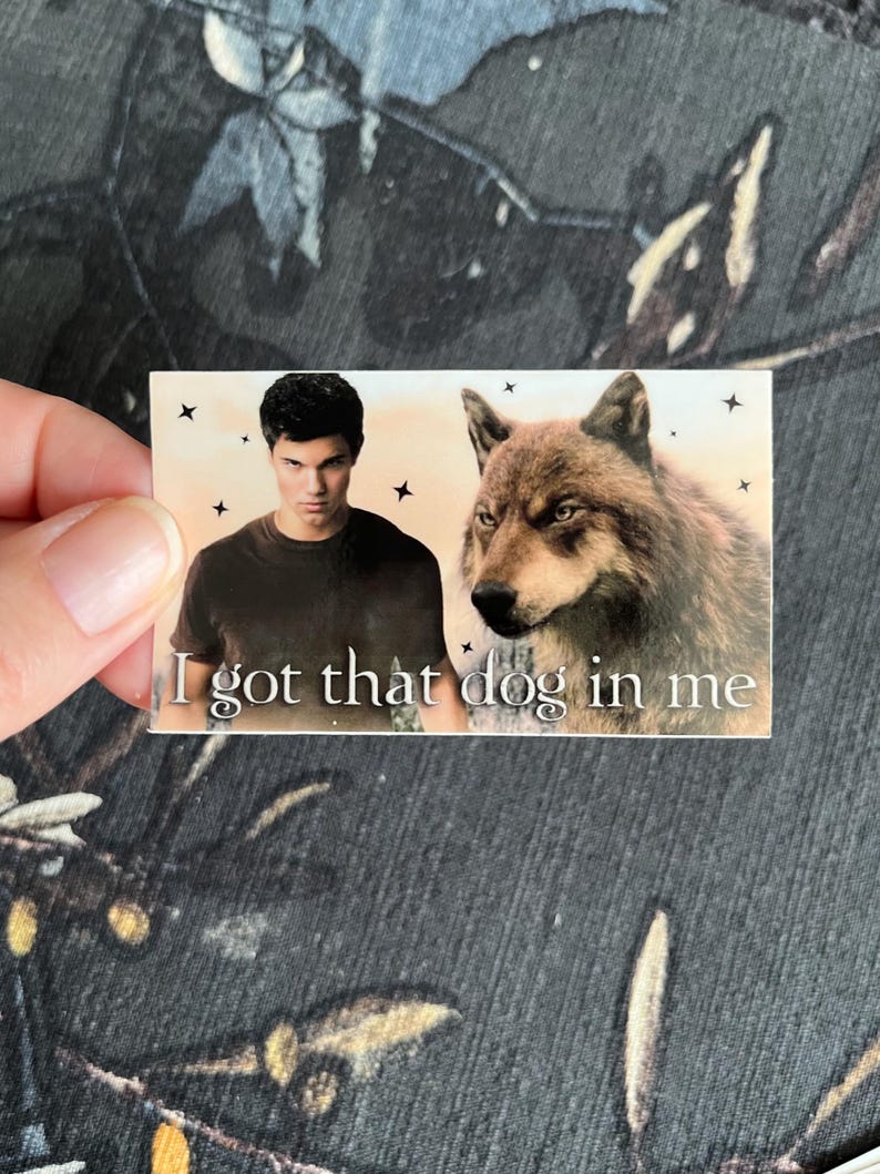 Op de afbeelding: Een rechthoekige sticker met een man en een wolf, met de tekst "I got that dog in me". De man draagt een donker shirt. De wolf heeft bruine vacht. De achtergrond is lichtoranje met zwarte sterren.