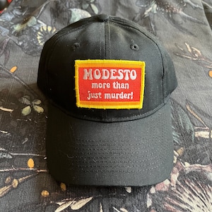 May include: Black baseball cap with a yellow and red patch that says "MODESTO more than just murder!"