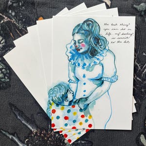 May include: A stack of white art cards featuring a watercolor illustration of two figures in clown-like attire. The figures are in shades of blue and white, with red accents. Text on the card reads, "the best thing you can do in life, my darling, is commit to the bit."