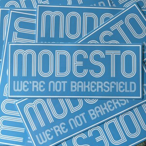 May include: A stack of blue and white stickers with the text "MODESTO WE'RE NOT BAKERSFIELD" in a retro font.