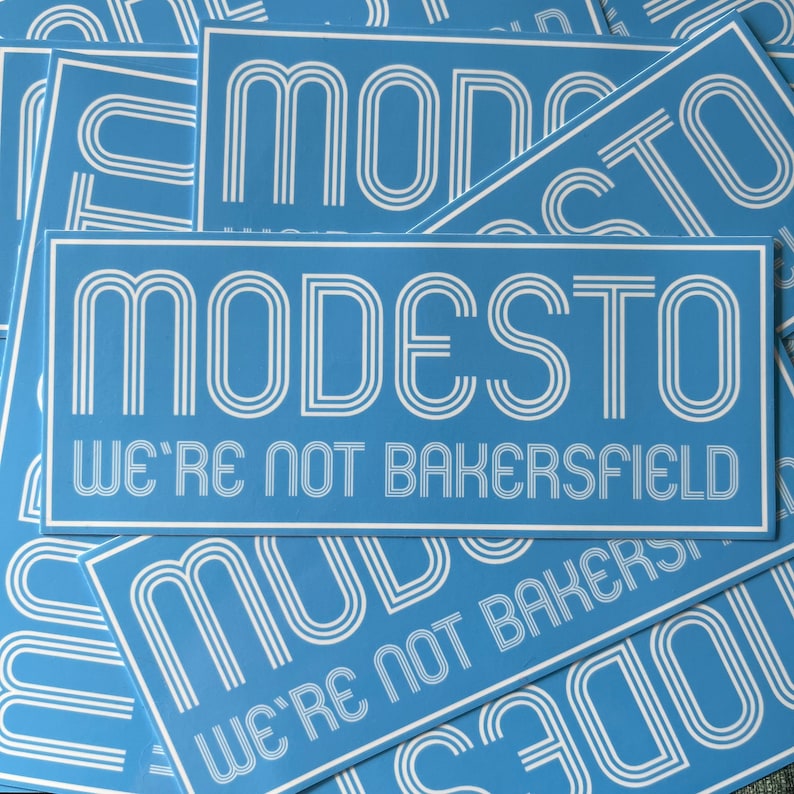 May include: A stack of blue and white stickers with the text "MODESTO WE'RE NOT BAKERSFIELD" in a retro font.