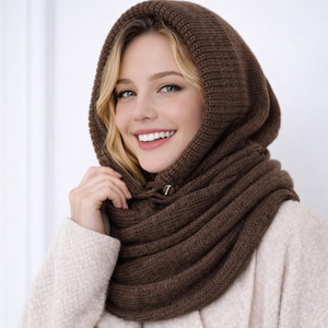 Può includere: Un cappuccio e sciarpa in maglia marrone. Il cappuccio è sollevato e la sciarpa avvolge il collo. Un bottone dorato fissa il cappuccio. Il materiale a maglia appare morbido e caldo, adatto per il clima freddo.