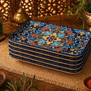 Puede incluir: Una pila de seis platos rectangulares de cerámica con un diseño floral vibrante. Los platos presentan un fondo azul oscuro con una flor central en amarillo y azul, rodeada de patrones de hojas naranjas y blancas. Los platos están apilados sobre una estera tejida.