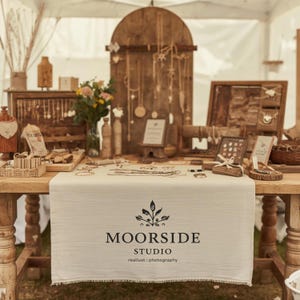 May include: A craft fair stall showcasing handmade jewellery. A cream linen tablecloth with the "Moorside Studio" logo covers the table. Necklaces, earrings, and other accessories are displayed on the table and a wooden backdrop, creating a rustic aesthetic.