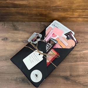 May include: A black gift box tied with twine, containing pink and red items, including a Hershey's milk chocolate bar, lipsticks, and a heart-shaped card. A tag reads "Rivals, Banter that flirts back, slow burn, Rom-Com Energy."