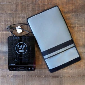May include: A black Westinghouse induction hob with a digital control panel and a circular heating zone, accompanied by a rectangular grey protective mat featuring black trim and two horizontal black straps, plus a black power cord with a plug.