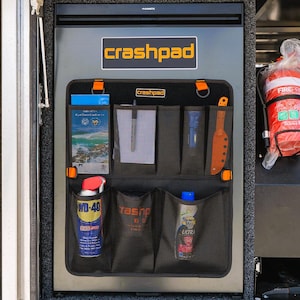 May include: A black Dometic refrigerator with a crashpad logo and a black fabric organiser attached to the front. The organiser features multiple mesh pockets holding a notebook, pen, torch, orange tool, WD-40 can, and sunscreen spray.