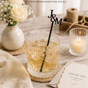 Puede incluir: Un vaso transparente con una bebida helada y un palito para remover con monograma negro que dice "J&M". La imagen también incluye un pequeño jarrón con flores blancas, una vela encendida y una tarjeta de agradecimiento. El texto "CUSTOM MONOGRAM STIR STICKS" está en la parte superior.