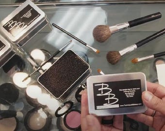 May include: An assortment of makeup brushes, compacts, and cleaning tools are arranged on a reflective surface. Several brushes with black handles and silver ferrules are visible, along with a black sponge and small metal containers with text. The overall theme is beauty and cosmetics.