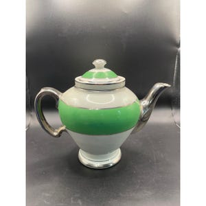 Pode incluir: Um bule de porcelana branca com uma larga faixa horizontal verde brilhante ao redor do centro e da tampa. O bule tem um acabamento prateado metálico na alça, no bico, no puxador da tampa e na base, com linhas prateadas finas.