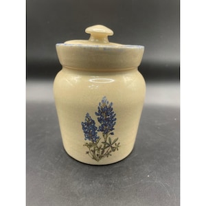 Puede incluir: Un tarro de cerámica beige con tapa, que presenta un diseño de flores de lupino pintado a mano en la parte frontal. El tarro tiene un acabado liso y moteado, y una tapa con borde azul que incluye un pequeño pomo como asa en la parte superior.