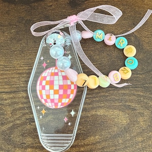 Puede incluir: Llavero acr&iacute;lico transparente con un dise&ntilde;o de bola de discoteca en rosa, naranja y blanco. El llavero tiene una pulsera de cuentas con la palabra "MIRROR" escrita con cuentas de colores. Se adjunta una cinta rosa.