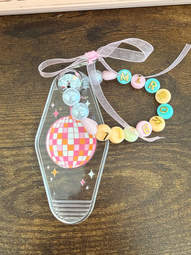 Puede incluir: Llavero acr&iacute;lico transparente con un dise&ntilde;o de bola de discoteca en rosa, naranja y blanco. El llavero tiene una pulsera de cuentas con la palabra "MIRROR" escrita con cuentas de colores. Se adjunta una cinta rosa.
