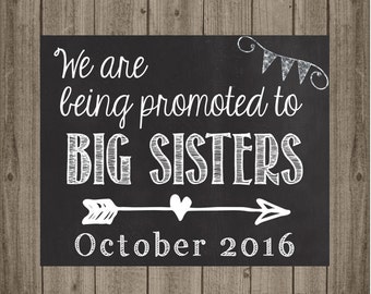 May include: Black chalkboard sign with white text that reads "We are being promoted to BIG SISTERS October 2016". There is a white arrow with a heart in the middle and a string of white flags above the text.