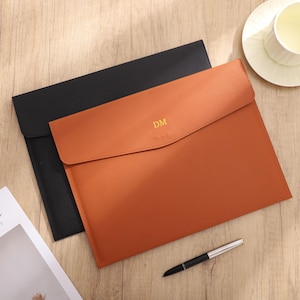 May include: Two leather document holders, one black and one tan, are displayed on a wooden surface. The tan holder has the initials "DM" in gold. A pen and a magazine with the word "ANTIC" are also visible.