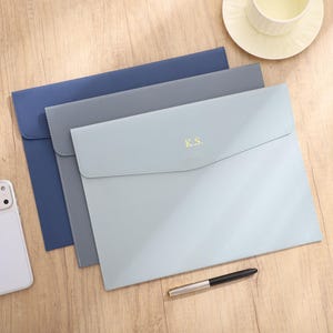 May include: Three leather document holders in blue and grey tones are arranged on a wooden surface. The front-most holder is light blue with the initials "K.S." in gold. A smartphone and pen are also visible.