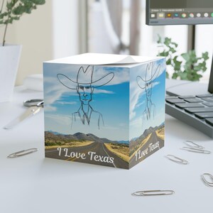 Puede incluir: Un cubo de bloc de notas blanco con un boceto de vaquero y el texto "I Love Texas". El diseño muestra una carretera a través de un paisaje desértico bajo un cielo azul. El bloc de notas está sobre una superficie blanca.
