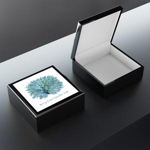 May include: Two black rectangular jewelry boxes, one open and one closed. The closed box features a peacock illustration in blue and green with the text "Keep Your Sparkle Safe." The open box reveals a white interior.