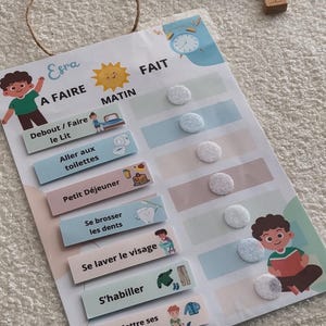 Peut inclure: Un tableau de routine matinale pour enfant avec le nom "Esra". Il comprend des tâches illustrées comme "Se lever/Faire le lit", "Aller aux toilettes" et "Petit Déjeuner". Une voiture jouet violette et d'autres jouets sont visibles.