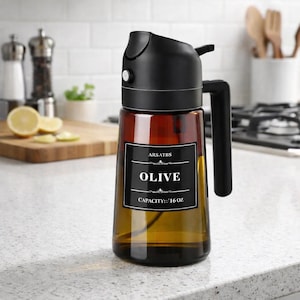 May include: A brown glass olive oil dispenser with a black handle and spout. The label reads "OLIVE" and "CAPACITY:: 16 OZ". The dispenser is on a white countertop in a kitchen setting.