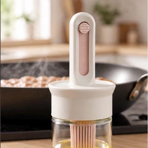Puede incluir: Un dispensador de aceite de vidrio transparente con tapa blanca y mango vertical que tiene un pincel de silicona rosa unido en la parte inferior. El mango incluye un botón deslizante rosa. El recipiente de vidrio contiene aceite amarillo.