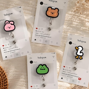 Puede incluir: Cuatro portatarjetas retráctiles con personajes de dibujos animados. Los personajes incluyen un conejito rosa, un oso marrón, una rana verde y un pato blanco. Cada portatarjetas está unido a una tarjeta blanca con un diseño estilo Instagram.