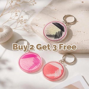 Op de afbeelding: Drie ronde sleutelhangers met zilveren sleutelringen. Elke sleutelhanger heeft een kleurrijk ontwerp met een roze, rode en gele gradiënt, en zwarte en witte lijnen. De tekst "Buy 2 Get 3 Free" wordt in de afbeelding weergegeven.