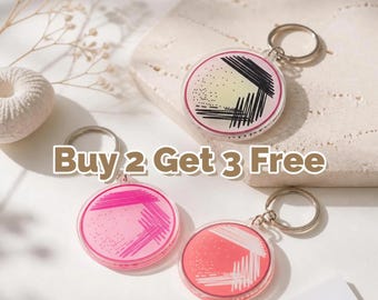 Puede incluir: Tres llaveros redondos con anillos plateados. Cada llavero presenta un diseño colorido con un degradado rosa, rojo y amarillo, y líneas negras y blancas. El texto "Buy 2 Get 3 Free" se muestra en la imagen.