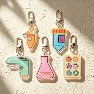 May include: A selection of vibrant acrylic keyrings with silver clasps. The designs feature a pen, a shield, a laboratory flask, a retro device, and a rectangle with circular patterns. The keyrings are displayed on a textured, light-coloured surface.