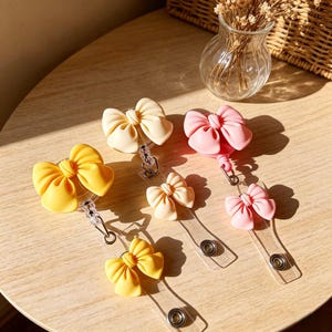 Può includere: Quattro porta badge retrattili con fiocchi decorativi in giallo, crema e rosa. Ogni porta badge ha un cinturino in plastica trasparente con un bottone a pressione in metallo. I fiocchi sono fissati all'alloggiamento circolare del meccanismo.