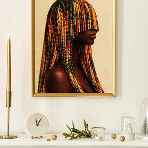 K&ouml;nnte beinhalten: Ein gerahmtes Kunstwerk zeigt eine Frau im Profil mit Perlenhaaren in Gelb-, Orange-, Gr&uuml;n- und Rott&ouml;nen. Die goldgerahmte Kunst h&auml;ngt &uuml;ber einem wei&szlig;en Kaminsims mit einer Uhr, einer Kerze und dekorativen Gegenst&auml;nden. Das Kunstwerk ist der Mittelpunkt.