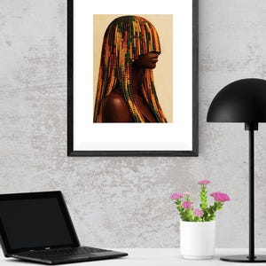 K&ouml;nnte beinhalten: Gerahmter Kunstdruck mit einem Portr&auml;t einer Person mit einer farbenfrohen Perlenfrisur. Das Kunstwerk befindet sich in einem schwarzen Rahmen und wird auf einer wei&szlig;en Oberfl&auml;che mit einem Laptop, einer kleinen wei&szlig;en Vase mit rosa Blumen und einer schwarzen Schreibtischlampe pr&auml;sentiert.