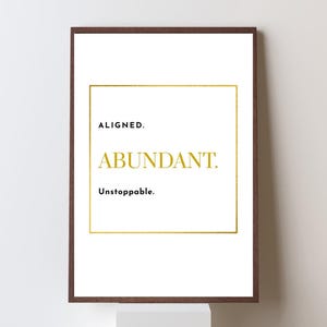 May include: A framed print with a white background and gold text. The words "ALIGNED.", "ABUNDANT.", and "Unstoppable." are displayed in a gold font. The frame is a dark brown color.