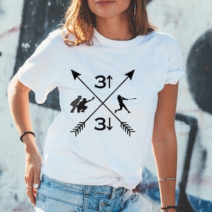 Può includere: Maglietta bianca con una grafica di baseball nera. La grafica presenta due giocatori di baseball, uno che batte e uno che riceve, con frecce che puntano verso l'alto e verso il basso e il numero 3.