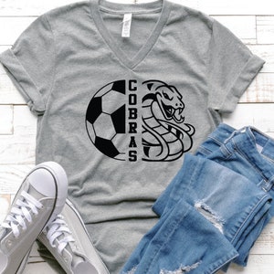 以下が含まれることがあります： グレーのVネックTシャツに、サッカーボールとコブラの白黒のグラフィックがプリントされています。Tシャツには「Cobras」の文字が書かれています。Tシャツは青いジーンズと合わせています。
