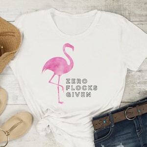 Puede incluir: Camiseta blanca con un gráfico de flamenco rosa y el texto "ZERO FLOCKS GIVEN". La camiseta está colocada sobre una superficie de madera con un sombrero de paja, sandalias y pantalones cortos de mezclilla.