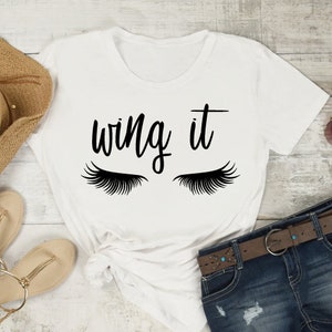 Puede incluir: Camiseta blanca con el texto "wing it" y un par de pestañas a cada lado del texto.