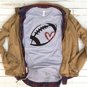 May include: A gray t-shirt with a black and white football graphic and a red heart. The shirt is folded and laid on a wooden surface with a brown jacket and a plaid shirt on top of it.