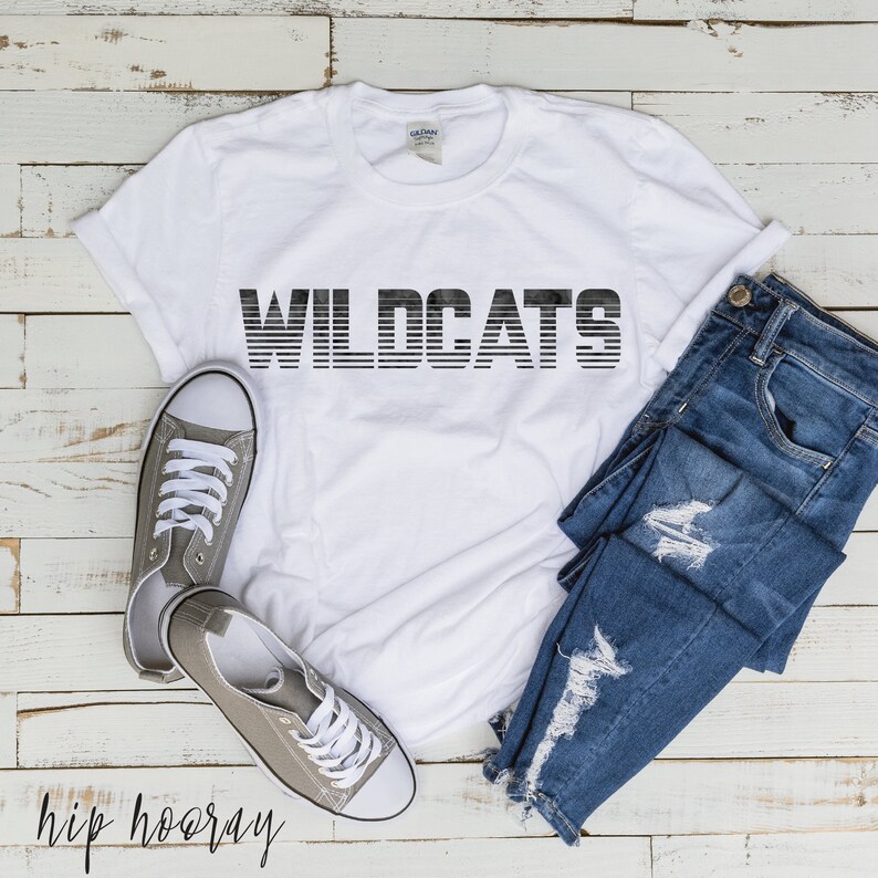 May include: White t-shirt with the word "WILDCATS" printed in black with a striped effect. The shirt is laid flat on a wooden surface with a pair of gray canvas shoes and blue jeans.