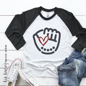 K&ouml;nnte beinhalten: Ein wei&szlig;-schwarzes Baseball-Raglan-Shirt mit einer grauen Baseballhandschuh-Grafik und einem roten Herzen darin. Das Shirt liegt auf einer wei&szlig;en Holzoberfl&auml;che mit einem Paar wei&szlig;en Turnschuhen und einer blauen Jeans.