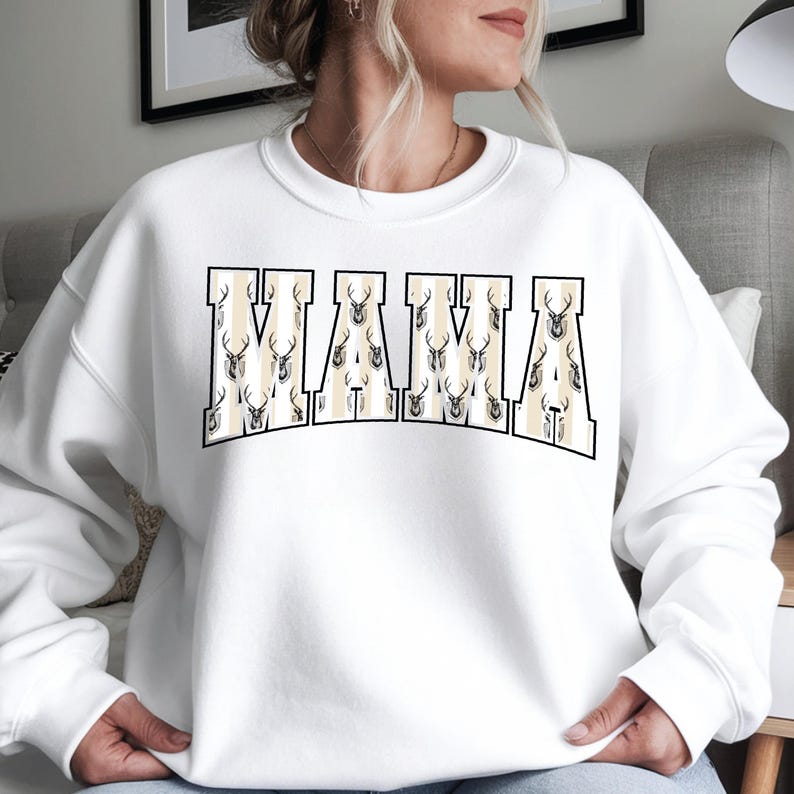 Może przedstawiać: Biała bluza z napisem "MAMA" w dużych, blokowych literach. Litery są beżowe ze wzorem poroża jelenia. Bluza w stylu casual, odpowiednia do noszenia na co dzień.