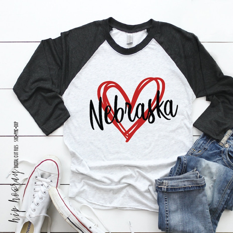 Peut inclure: Un t-shirt de baseball raglan avec un corps blanc et des manches gris fonc&eacute;. Le t-shirt pr&eacute;sente un c&oelig;ur rouge et le mot "Nebraska" en &eacute;criture noire. Le tout est accompagn&eacute; d'un jean bleu et de baskets blanches.