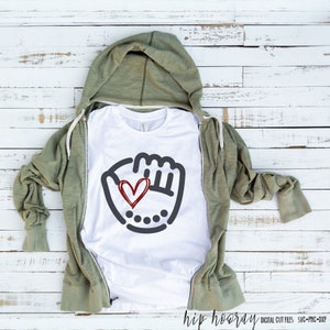 K&ouml;nnte beinhalten: Wei&szlig;es T-Shirt mit einem grauen Baseballhandschuh-Motiv und einem roten Herzen darin. Das Shirt ist gefaltet und auf einer wei&szlig;en Holzoberfl&auml;che abgelegt.