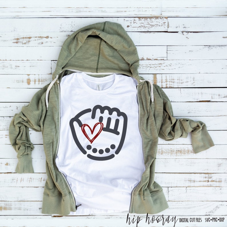 K&ouml;nnte beinhalten: Wei&szlig;es T-Shirt mit einem grauen Baseballhandschuh-Motiv und einem roten Herzen darin. Das Shirt ist gefaltet und auf einer wei&szlig;en Holzoberfl&auml;che abgelegt.