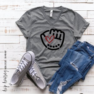 K&ouml;nnte beinhalten: Graues V-Ausschnitt-T-Shirt mit einem schwarzen und roten Baseballhandschuh-Motiv mit einem Herzen darin. Das Shirt liegt auf einer wei&szlig;en Holzoberfl&auml;che mit einem Paar grauen Canvas-Schuhen und einer blauen Jeans.