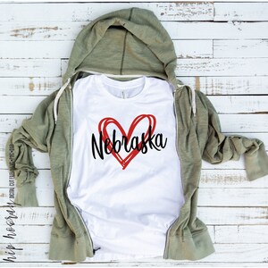 Peut inclure: T-shirt blanc avec un c&oelig;ur rouge et le mot "Nebraska" en noir. Le t-shirt est pr&eacute;sent&eacute; sur un sweat &agrave; capuche vert zipp&eacute;. Le fond est constitu&eacute; de planches de bois blanches. Fichier de d&eacute;coupe num&eacute;rique.