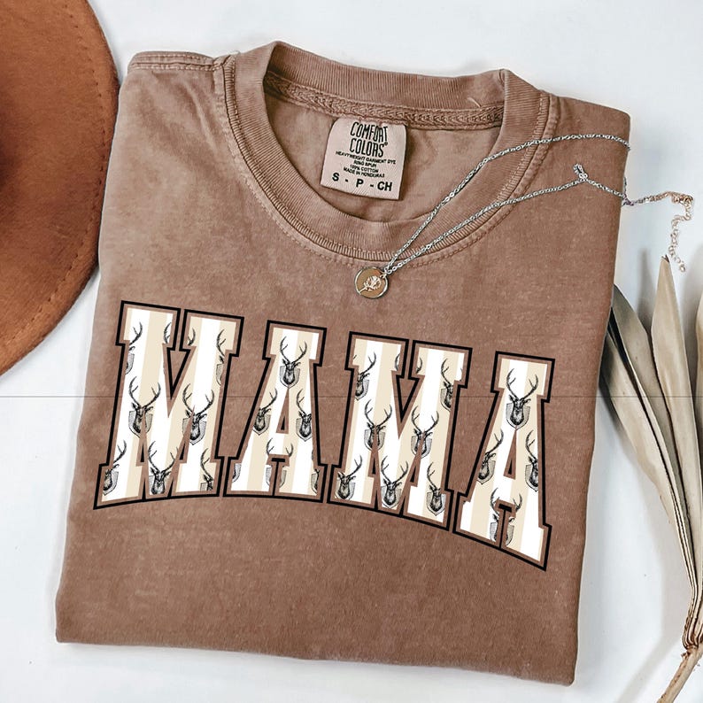Może przedstawiać: Brązowa koszulka Comfort Colors z napisem "MAMA" w stylu uniwersyteckim. Litery wypełnione są wzorem poroża jelenia. Srebrny naszyjnik z wisiorkiem jest ułożony na koszulce.