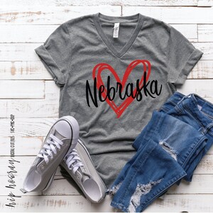 Peut inclure: Un t-shirt gris &agrave; col en V avec un c&oelig;ur rouge et le mot "Nebraska" en &eacute;criture noire. L'image pr&eacute;sente &eacute;galement un jean bleu et des baskets grises &agrave; lacets blancs. L'ensemble sugg&egrave;re un style d&eacute;contract&eacute;.